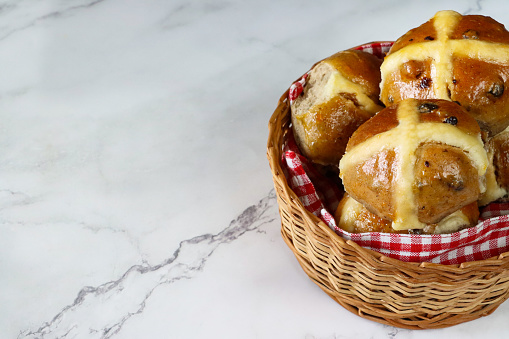 Stock photo showing close-up view of a pile of freshly baked, homemade Easter hot cross buns in a wicker basket lined with a red and white gingham cloth, home baking concept.\nThese traditional spiced sweet buns are made and sold over the Easter period, with the cross symbol on the glazed top being made from a flour and water paste, and symbolising Christianity.