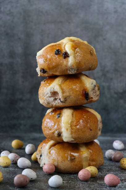 Stock photo showing close-up view of stack display of freshly baked, homemade Easter hot cross buns, home baking concept. These traditional spiced sweet buns are made and sold over the Easter period, with the cross symbol on the glazed top being made from a flour and water paste, and symbolising Christianity. imagen en primer plano de una pila de bollos de cruz calientes de pascua recién horneados y caseros rodeados de mini huevos de pascua de chocolate recubiertos de caramelos moteados, fondo gris moteado, concepto de horneado casero, enfoque en primer plano