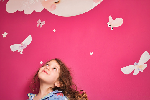 Young cute girl wearing pajamas looking up at a pink wall with decal stars, moon and fairies