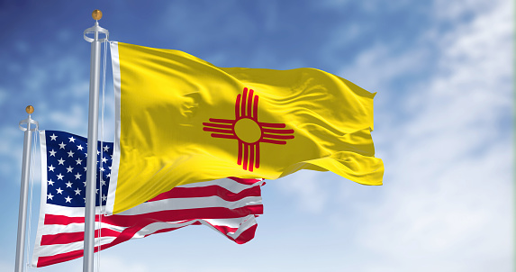 The New Mexico state flag waving along with the national flag of the United States of America. In the background there is a clear sky. New Mexico is a state in the Southwestern United States