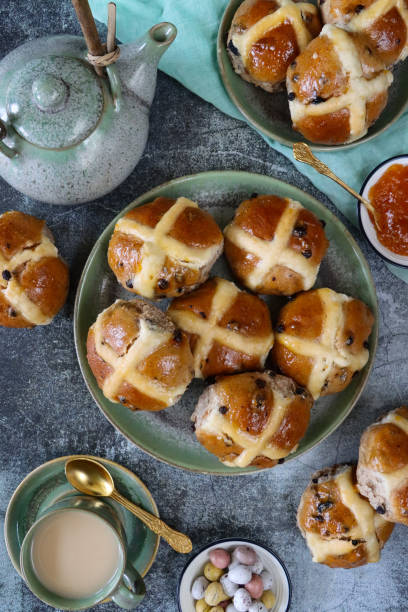 Stock photo showing elevated view of china teapot besides plates containing freshly baked, homemade Easter hot cross buns, home baking concept.
These traditional spiced sweet buns are made and sold over the Easter period, with the cross symbol on the glazed top being made from a flour and water paste, and symbolising Christianity. imagen de bollos cruzados calientes caseros de pascua en platos además de tetera de porcelana y taza de té en platillo con cucharadita, platos de mermelada de albaricoque y mini huevos de chocolate, muselina verde, fondo gris moteado, vista elevada