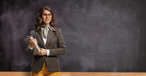 Young female teacher holding books and posing in front of a chalkboard