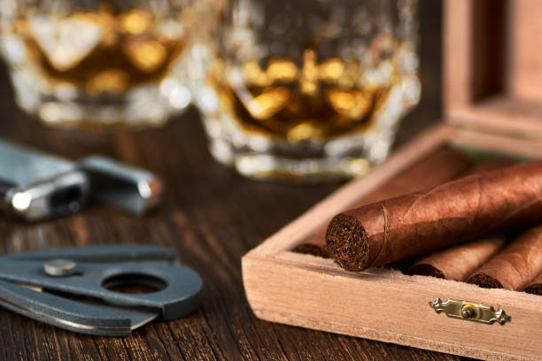 vista di una scatola di legno con sigari cubani, un accendino e un taglierino. natura morta con due bicchieri di whisky sul tavolo. - cigar whisky bar cognac foto e immagini stock
