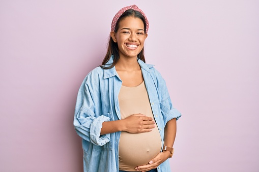Beautiful hispanic woman expecting a baby, touching pregnant belly smiling and laughing hard out loud because funny crazy joke.
