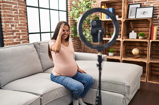 Young pregnant woman recording vlog tutorial with smartphone at home smiling happy doing ok sign with hand on eye looking through fingers