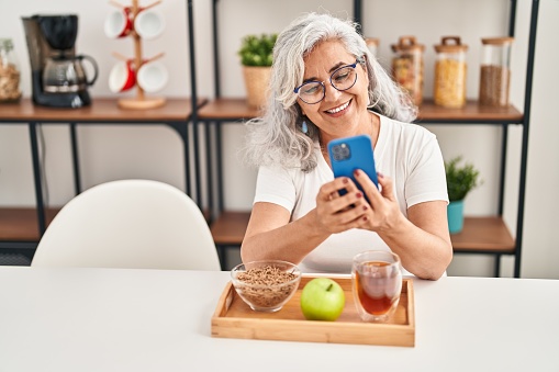 Middle age woman having breakfast using smartphone at home