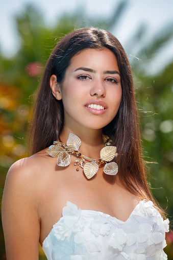 Beautiful young woman with long brown hair in white strapless top wearing leafy necklace, standing in public park and smiling at camera, beauty and fashion industry