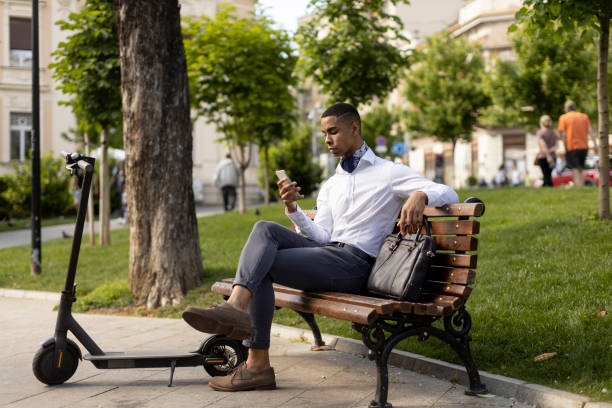 istock Young African American using mobile phone while sitting on the bench by electric scooter 1396253989