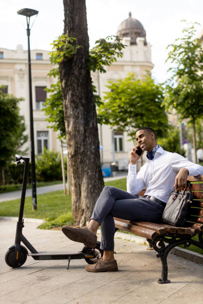 istock Young African American using mobile phone while sitting on the bench by electric scooter 1396254000