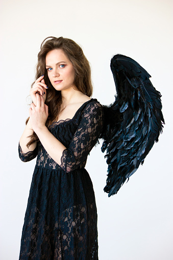 Pretty young brunette woman (European, American female) wears black dress and black wings on white background, vertical plane, studio photo. Halloween, parade, festival concept.