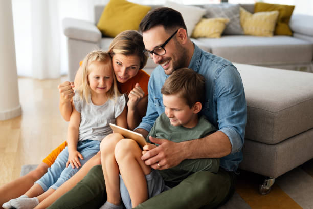 счастливая молодая семья весело проводит время дома. родители с детьми используют цифровое устройство. - ipad iphone smart phone ipad 3 стоковые фото и изображения