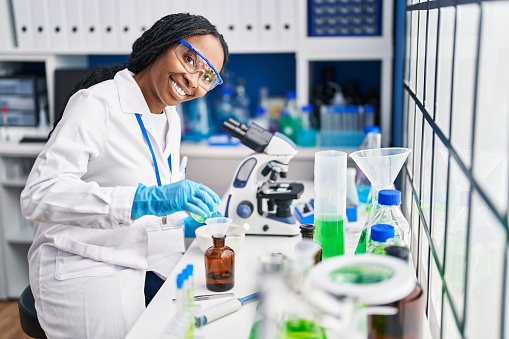 African american woman wearing scientist uniform pouring liquid on bottle laboratory
