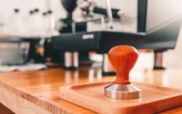 Close-up of a barista's tool Coffee Tamper with Wooden Handle placed on a wooden bar counter with a coffee machine in a coffee shop.