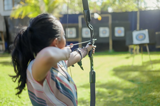 Rearview shot of an Indonesian female archer stretching her bow while aiming it at a target