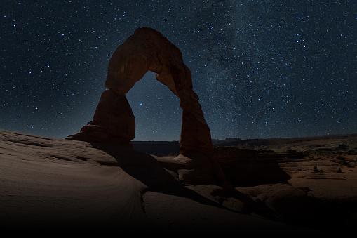 The Pack Creek Fire in the La Sal Mountains lights up the night sky in the distance, with the Milky Way over Delicate Arch in Arches National Park, Utah.