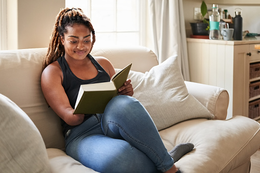 Young African woman reading a book while sitting on living room sofa at home, woman missing lower half of her arm