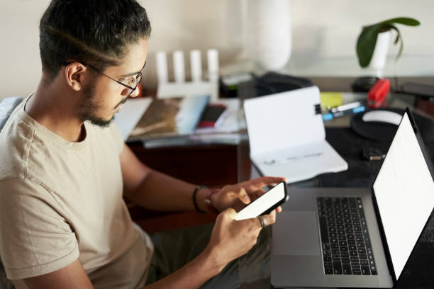 istock Young man sitting at table with laptop using phone at home 1396282486