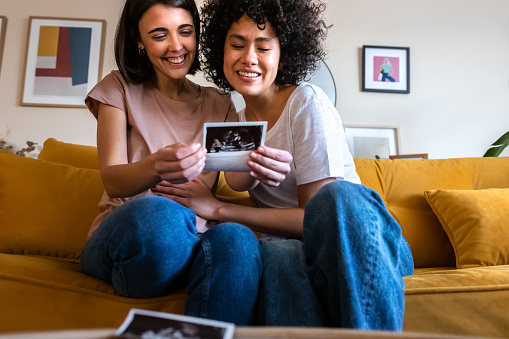 Smiling multiracial lesbian pregnant couple looking baby ultrasounds at home sitting on the couch. LGTBQ love and family concept.