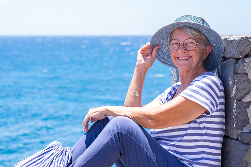 Happy senior woman in striped t-shirt sitting by the sea holding her hat lest it fly away. Mature lady dressed in blue enjoying relaxation and vacation looking at camera