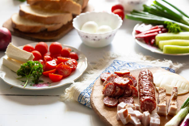 A traditional Romanian lunch with plates of fresh vegetables - spring onions, tomatoes, cucumbers. There is fresh meat including salami, sausage and slanina - cubes of pork fat. There are hard boiled eggs and a loaf of fresh bread. All of this is presented on a white wooden surface.