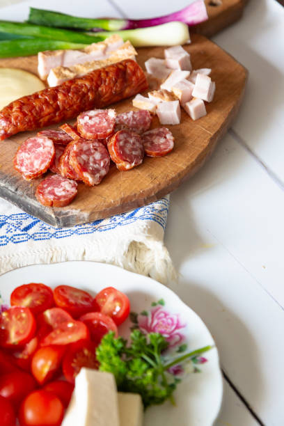 A traditional Romanian lunch with plates of fresh vegetables - spring onions, tomatoes, cucumbers. There is fresh meat including salami, sausage and slanina - cubes of pork fat. There are hard boiled eggs and a loaf of fresh bread. All of this is presented on a white wooden surface.