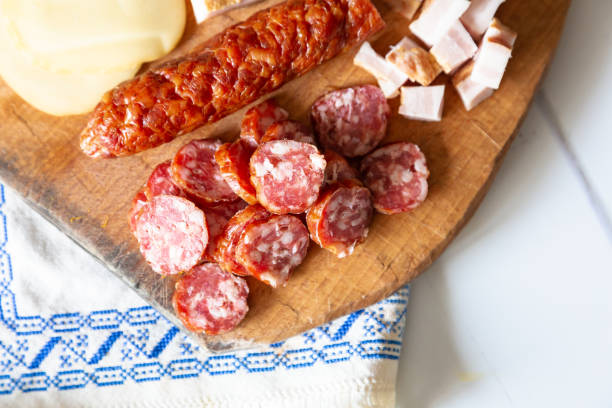 A traditional Romanian lunch with plates of fresh vegetables - spring onions, tomatoes, cucumbers. There is fresh meat including salami, sausage and slanina - cubes of pork fat. There are hard boiled eggs and a loaf of fresh bread. All of this is presented on a white wooden surface.