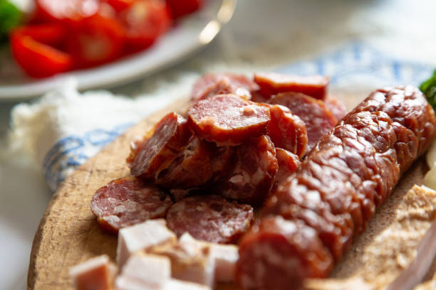A traditional Romanian lunch with plates of fresh vegetables - spring onions, tomatoes, cucumbers. There is fresh meat including salami, sausage and slanina - cubes of pork fat. There are hard boiled eggs and a loaf of fresh bread. All of this is presented on a white wooden surface.