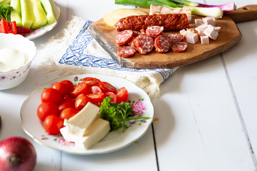 A traditional Romanian lunch with plates of fresh vegetables - spring onions, tomatoes, cucumbers. There is fresh meat including salami, sausage and slanina - cubes of pork fat. There are hard boiled eggs and a loaf of fresh bread. All of this is presented on a white wooden surface.