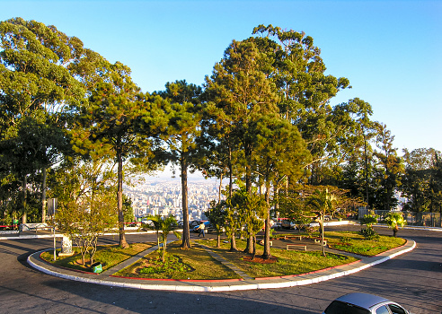 Viewpoint in a square with blue sky, trees, buildings, favela, avenues and buildings. In the background, the city of Belo Horizonte, framed by trees.