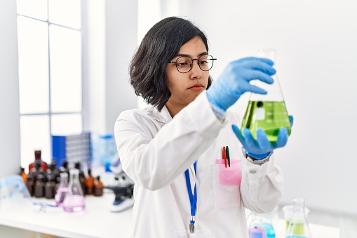 Young latin woman wearing scientist uniform measuring liquid on test tube at laboratory