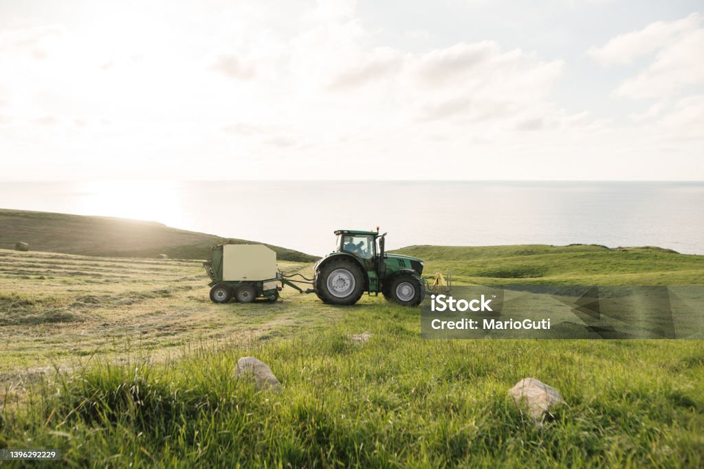 Tractor with baler in a grass field Tractor with baler in a grass field by the sea at sunset Hay Baler Stock Photo