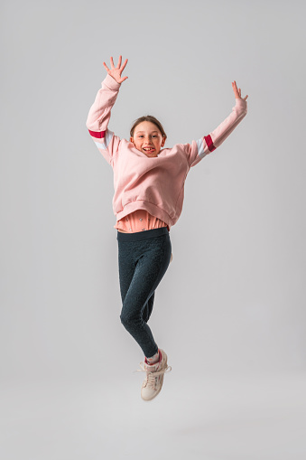 Portrait of a Caucasian teenage girl in the studio. She is jumping in the air with her arms raised. She is looking at the camera. White background.