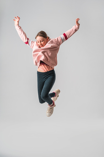 Portrait of a Caucasian teenage girl in the studio. She is jumping in the air with her arms raised. She is looking down. White background.