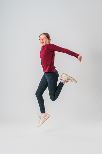 Portrait of a Caucasian teenage girl in the studio. She is jumping in the air with her arms raised. She is looking at the camera. White background.