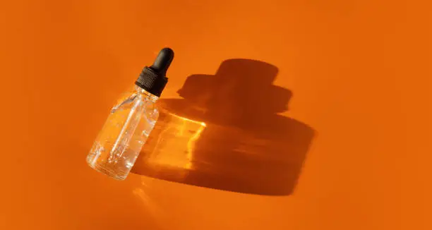 Transparent glass dropper bottle with air bubbles on orange background in sunlight. Pipette with fluid hyaluronic acid, serum, retinol. Cosmetics and healthcare concept Flat lay. Luxury beauty product
