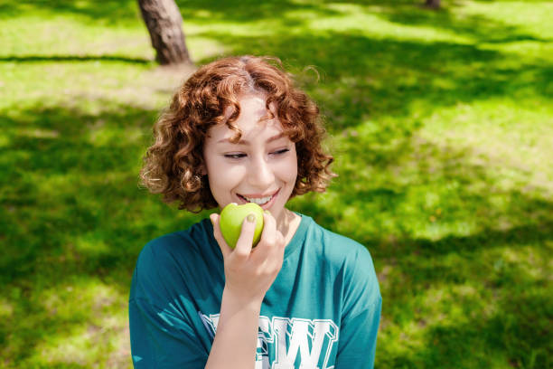 Portrait young charming redhead woman wearing green tee biting green apple with big smiles while looking away background summer park. stock photo