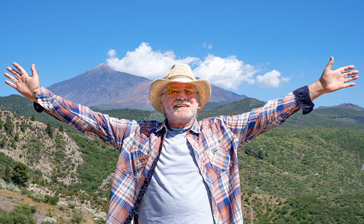 Bearded senior man with straw hat standing outdoors enjoying hike. Mountains with the Teide volcano in the background