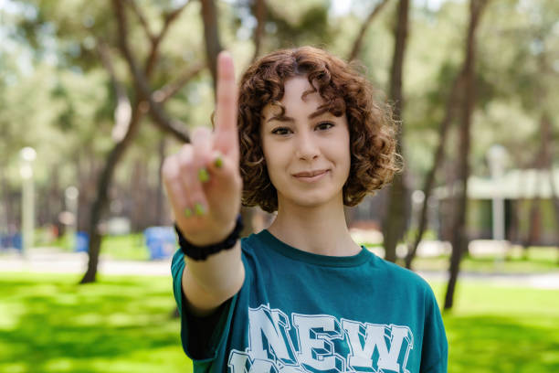 Young redhead woman wearing green tee showing number one with finger with a bit of smile, outdoors. Selective focus on her face. stock photo