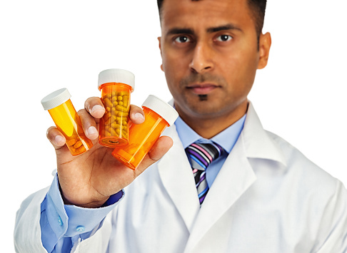 Photo of a serious Indian pharmacist showing a group of three prescription pill bottles in one hand; isolated on white. Primary focus on bottles and hand in foreground.