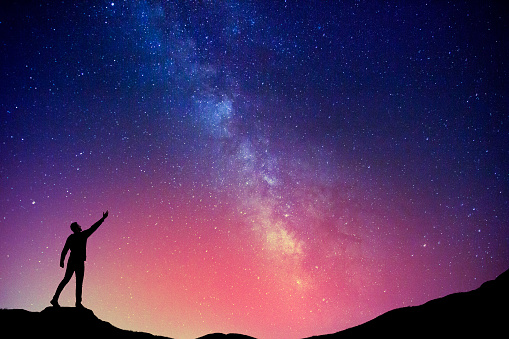 Man standing on the cliff with one hand up admiring the beautiful sky with Milky Way.