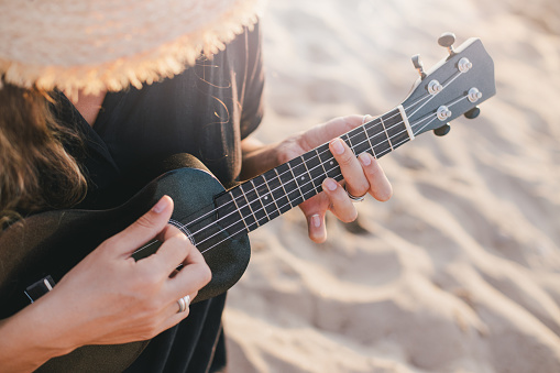 Beautiful young woman with long curly healthy hair wearing trendy straw hat playing ukulele at the beach.