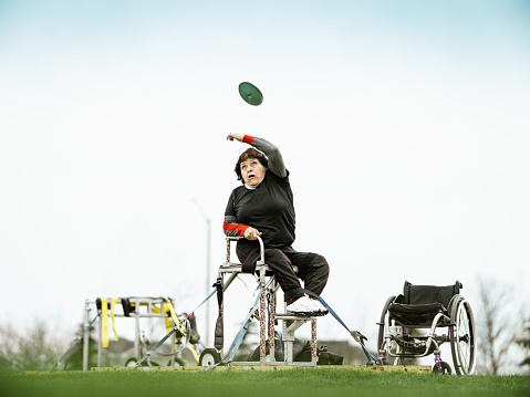 Senior Latin disabled lady practising discus throwing. She is dressed in sport outfit. Exterior of public park sport field during cloudy day.
