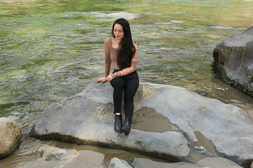 A Chinese model sitting on a rock beside a river in Spring. She is wearing a brown top, black pants and boots.