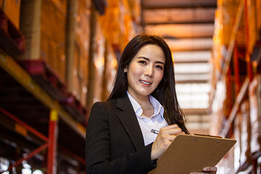 Portrait of Asian business woman or vice president smiling and holding clipboard at warehouse factory. Confidence people with positive vision working at import export company concept with copy space