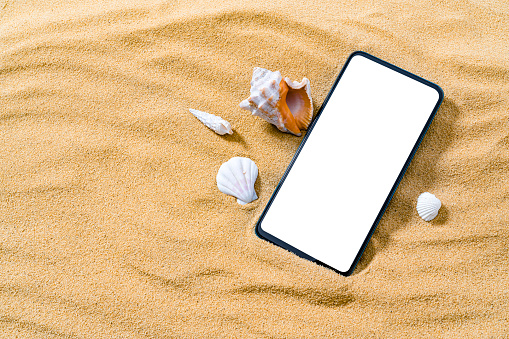 Summer backgrounds: high angle view of a smartphone with blank screen laying on golden beach sand background. Conch shells complete the composition. Copy space on blank screen. File contain path on mobile screen. High resolution 42Mp studio digital capture taken with SONY A7rII and Zeiss Batis 40mm F2.0 CF lens