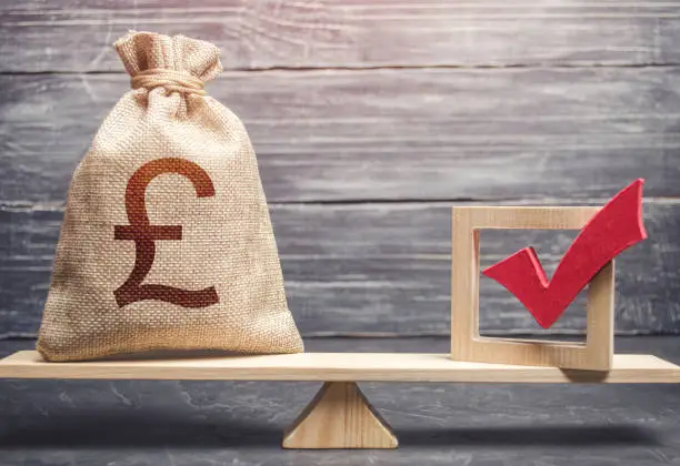 Photo of Red vote tick and a british pound sterling money bag on scales. Estimating cost of making a decision and consequences in the future. Corruption risks. Concept of lobbying for decisions and laws.