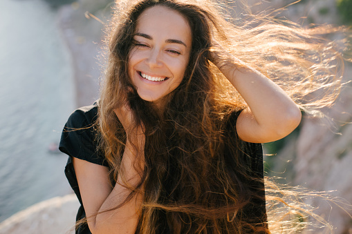 Young beautiful happy woman with long curly hair posing on a top of the mountain with sea view.