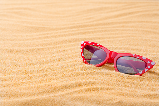 Red female sunglasses on golden tropical sandy beach. The composition is at the right of an horizontal frame leaving useful copy space for text and/or logo at the left. High resolution 42Mp outdoors digital capture taken with SONY A7rII and Zeiss Batis 40mm F2.0 CF lens