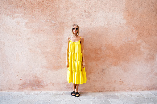 Fashionable blonde woman in yellow summer dress and sunglasses standing in front of the wall. A lot of copy space.