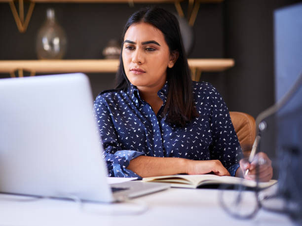 istock Focused indian businesswoman writing in a notebook while working alone on a laptop in an office. One female only making notes in her diary and planning online while working at her desk 1396321415